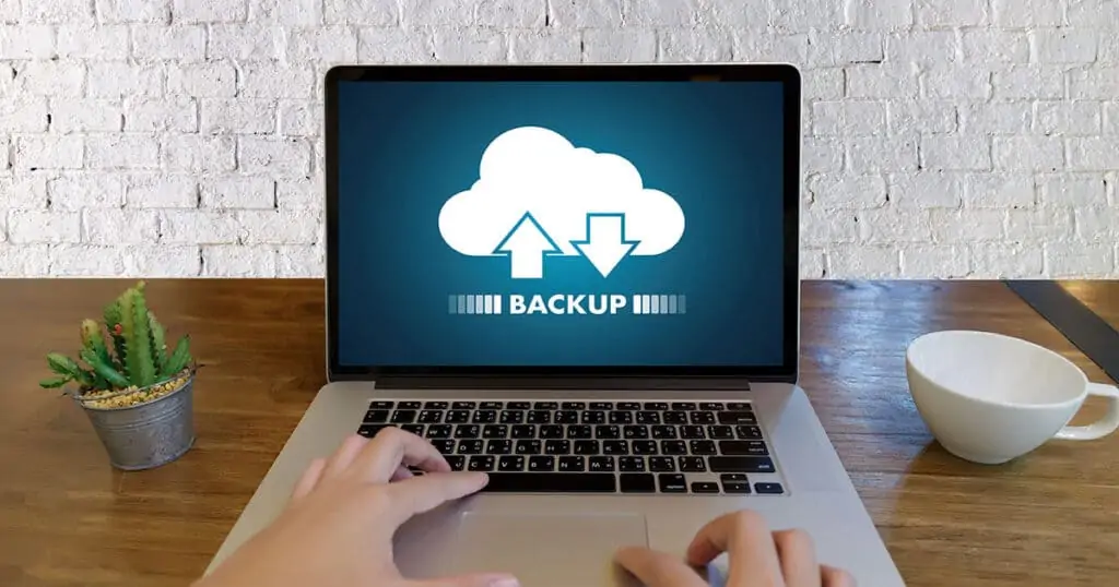 A laptop on a wooden desk displays a backup icon, with a person's hands on the keyboard, a white cup, and a plant beside it.