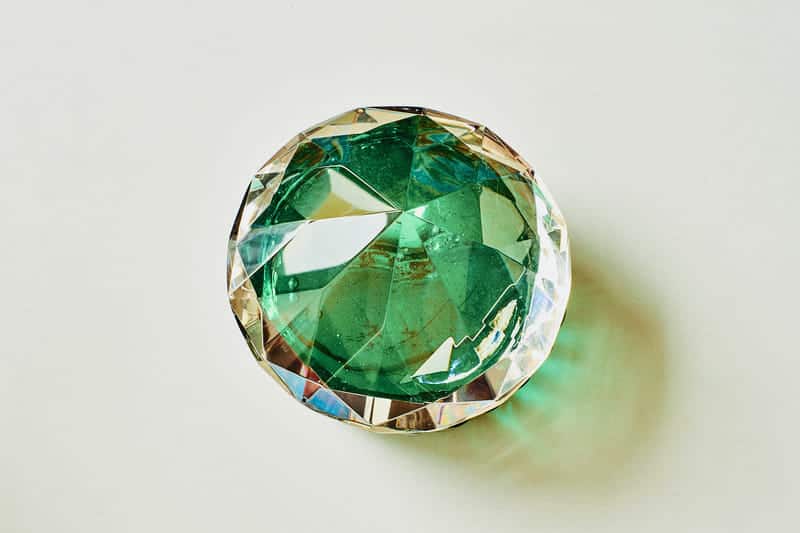 A large, multifaceted gemstone with variations in color and clarity, resting on a light surface with a shadow cast to the side.